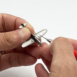 Puede incluir: Un pequeño modelo de avión plateado se sostiene entre dos manos. Se utiliza una herramienta Eiger roja y blanca para trabajar en el modelo. El avión tiene un diseño detallado, incluyendo alas y cola. El fondo es blanco.