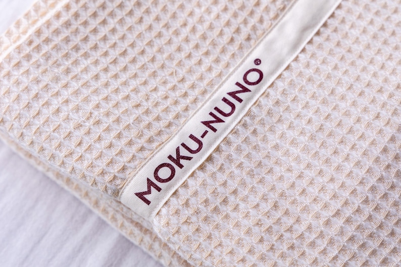 May include: A close-up of a beige waffle weave towel with a white label that says "MOKU-NUNO" in burgundy lettering.