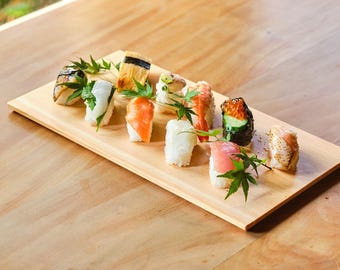 May include: A wooden serving board displays an assortment of sushi. The sushi includes salmon, tuna, shrimp, and other seafood varieties, garnished with green leaves. The board rests on a light-colored wooden surface.