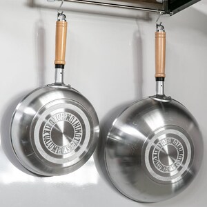 Pu&ograve; includere: Due wok da cucina argentati con manici in legno appesi a un rack di metallo. I wok hanno un design circolare sul fondo con il testo "CAST ALUMINUM WOK"