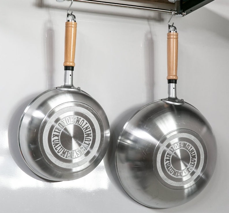 Pu&ograve; includere: Due wok da cucina argentati con manici in legno appesi a un rack di metallo. I wok hanno un design circolare sul fondo con il testo "CAST ALUMINUM WOK"