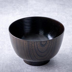 May include: A dark brown wooden bowl with a smooth, polished interior and a textured exterior. The bowl has a rounded shape with a slightly angled rim and a small, circular base. The wood grain is visible, adding to its natural aesthetic.
