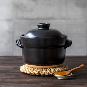 May include: A black ceramic cooking pot with a lid, set on a woven trivet. A small wooden spoon rests on a white saucer. The pot has two handles and is on a dark wooden surface.