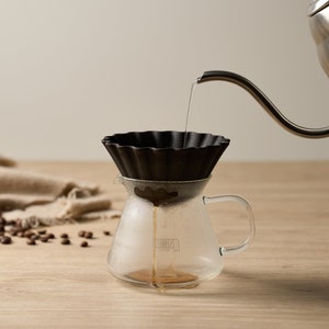 May include: A black pour-over coffee filter sits in a clear glass carafe with a handle. Water is being poured from a silver kettle into the filter.