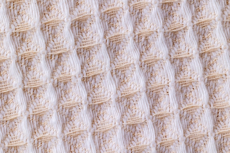 May include: Close-up of a white and beige waffle weave fabric. The fabric has a textured, raised pattern.
