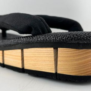 May include: Black fabric sandals with a wooden platform sole. The sole is made of multiple pieces of wood, creating a layered effect.