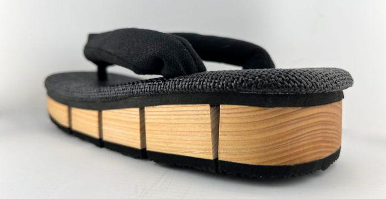 May include: Black fabric sandals with a wooden platform sole. The sole is made of multiple pieces of wood, creating a layered effect.