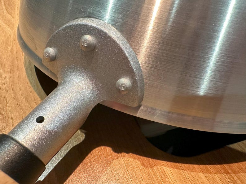 Pu&ograve; includere: Primo piano di un manico di wok in metallo argentato con quattro rivetti. Il manico &egrave; attaccato al wok con un pezzo di metallo curvo.