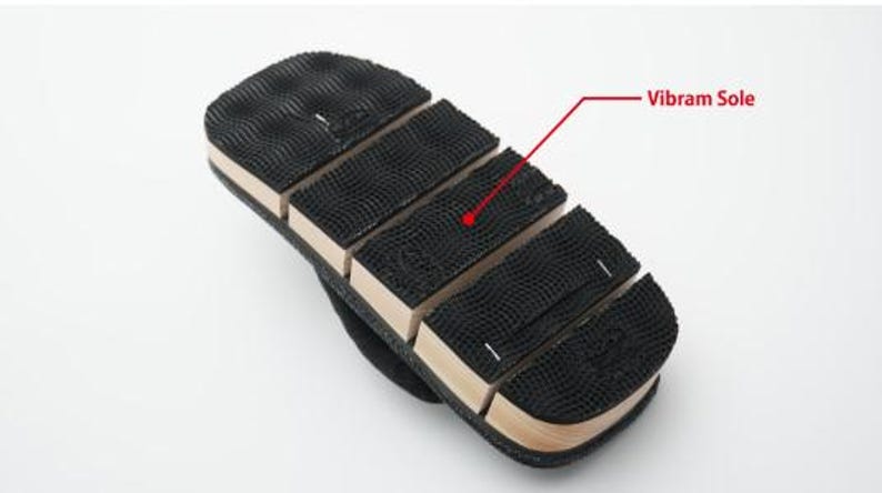 May include: Close-up of the bottom of a black sandal with a brown wooden base. The sole is made of black rubber with a textured pattern and the text "Vibram Sole" is written in red.