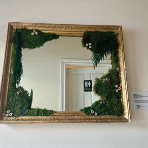 May include: A rectangular mirror with a gold frame, decorated with green moss and small white flowers. The mirror reflects a white door with a restroom sign. The frame has an ornate, textured design.