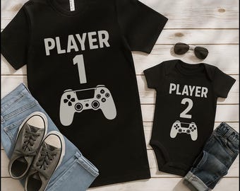 May include: Black t-shirt with "PLAYER 1" and a game controller graphic, paired with a matching black onesie with "PLAYER 2" and a controller. Includes jeans, sunglasses, and gray sneakers.