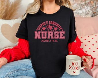 以下が含まれることがあります： ピンクのグラフィックがプリントされた黒いTシャツ。グラフィックには「Cupid's Favorite Nurse」の文字と、ピンクのハートとキューピッドのデザインが描かれています。グラフィックの下には「Ashley R.N.」という名前がプリントされています。