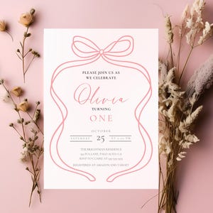 Op de afbeelding: Een roze en witte uitnodiging voor een eerste verjaardag. De uitnodiging heeft een roze lintontwerp rond de tekst "Olivia Turning One". Details zijn de datum, tijd en locatie van het feest.