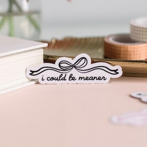 May include: A white sticker featuring a black bow design and the text "i could be meaner". The sticker is set against a light pink surface, with a white book and rolls of decorative tape visible.