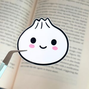 May include: A white sticker in the shape of a dumpling with a smiling face, black eyes, and pink cheeks. The sticker is held by a pair of tweezers against the backdrop of an open book.