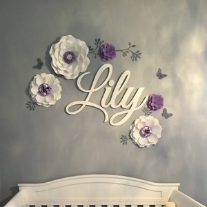 May include: A wall decoration featuring the name "Lily" in cursive script, surrounded by large paper flowers in white and purple, with gray butterfly accents. The flowers have detailed centers and are arranged on a gray wall.