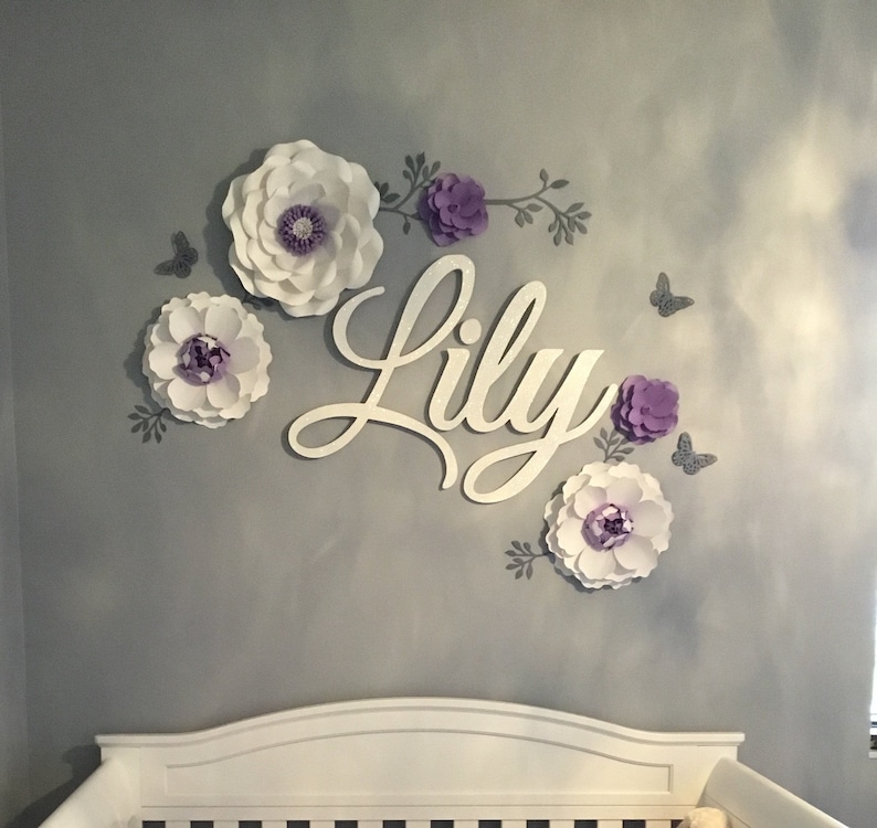May include: A wall decoration featuring the name "Lily" in cursive script, surrounded by large paper flowers in white and purple, with gray butterfly accents. The flowers have detailed centers and are arranged on a gray wall.