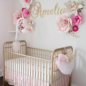 May include: A gold-colored crib with a pink ruffled skirt and a white and gray patterned sheet. The wall is decorated with large paper flowers in shades of pink and cream, and the name "Amelia" in gold script.