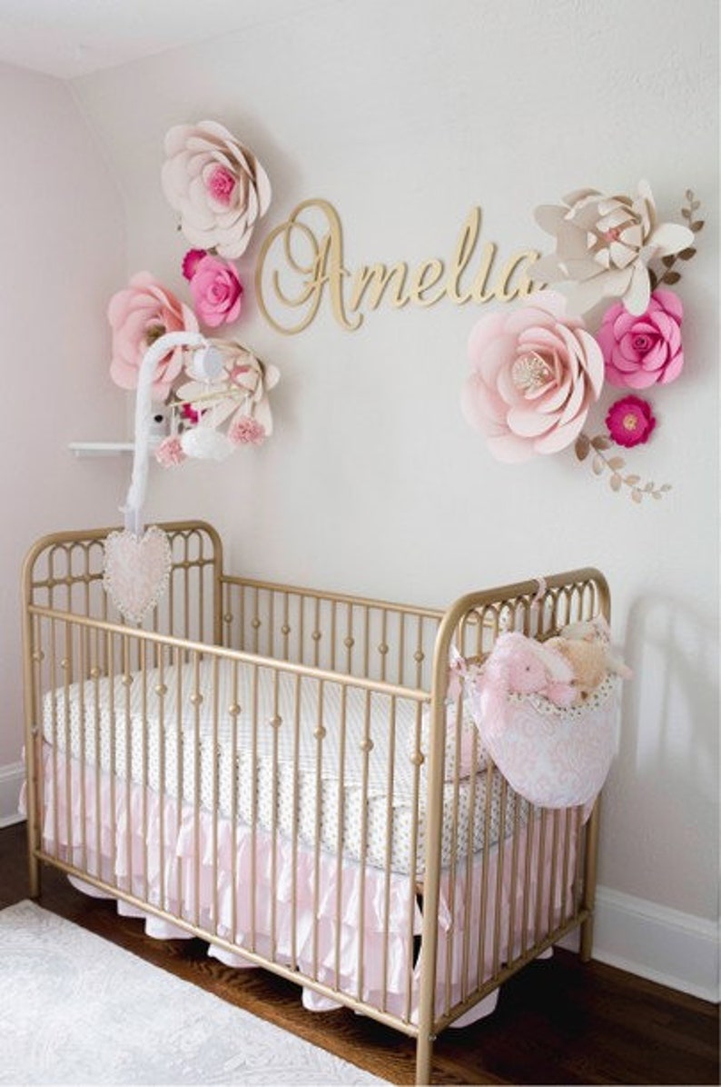 May include: A gold-colored crib with a pink ruffled skirt and a white and gray patterned sheet. The wall is decorated with large paper flowers in shades of pink and cream, and the name "Amelia" in gold script.