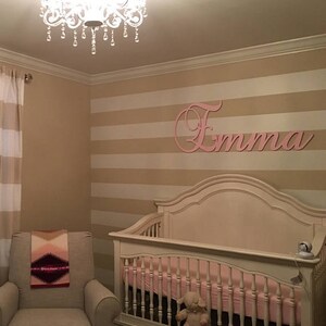 May include: A nursery with a crib, a rocking chair, and a decorative chandelier. The walls are painted with horizontal stripes. A pink wooden name, "Emma", is mounted on the wall above the crib. A blanket is draped over the chair.