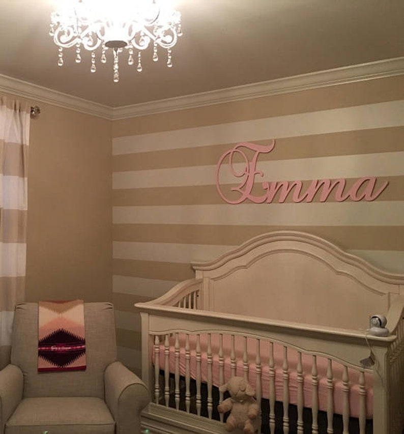 May include: A nursery with a crib, a rocking chair, and a decorative chandelier. The walls are painted with horizontal stripes. A pink wooden name, "Emma", is mounted on the wall above the crib. A blanket is draped over the chair.