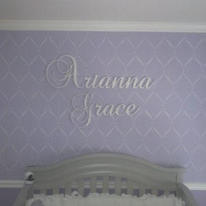 May include: White cursive name sign, "Arianna Grace", mounted on a lavender wall with a diamond pattern. A gray crib is visible in the foreground, suggesting a nursery setting.