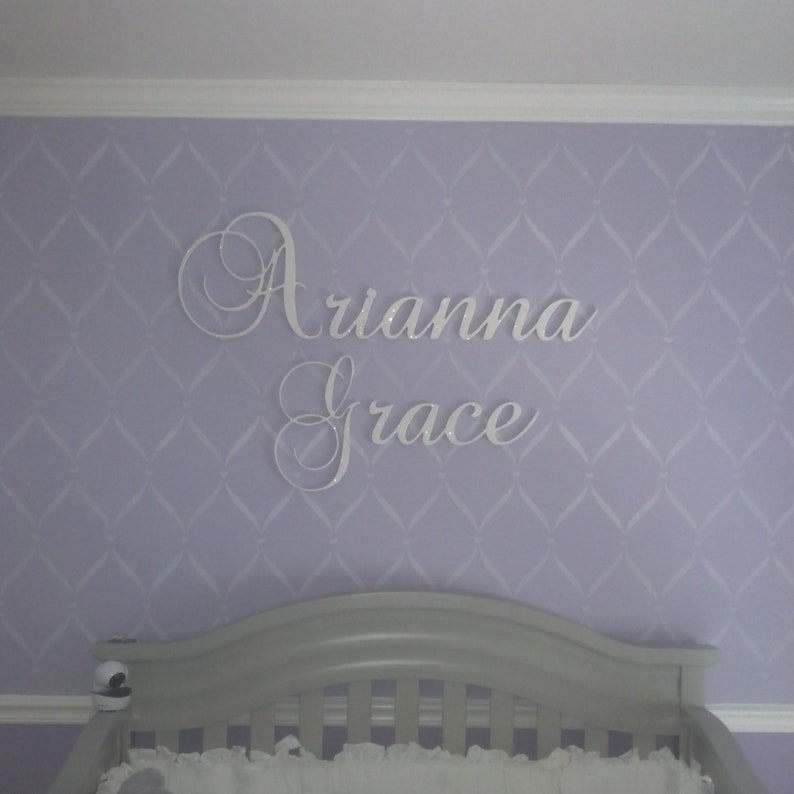 May include: White cursive name sign, "Arianna Grace", mounted on a lavender wall with a diamond pattern. A gray crib is visible in the foreground, suggesting a nursery setting.