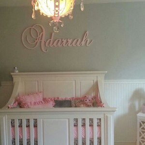 May include: A white crib with pink bedding and a matching rug. A pink chandelier hangs above the crib, and the name "Adarrah" is displayed on the wall. The room has a soft, feminine aesthetic.