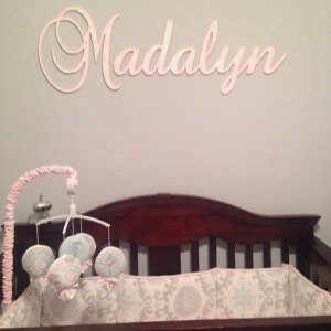 May include: A light pink wooden sign that spells out the name "Madalyn" in cursive script is mounted on a light gray wall. Below the sign is a dark brown crib with a patterned bumper and a mobile with hanging plush toys.