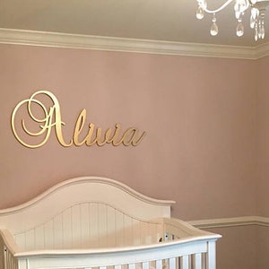 May include: A nursery with a white crib and a gold-colored name sign that reads "Olivia" on the wall. Two framed pictures hang on the wall. The room is painted a light pink color.