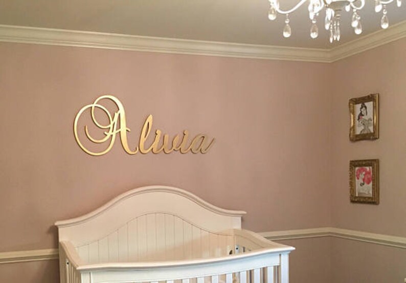 May include: A nursery with a white crib and a gold-colored name sign that reads "Olivia" on the wall. Two framed pictures hang on the wall. The room is painted a light pink color.
