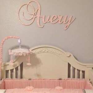 May include: A nursery scene featuring a crib with pink bedding and a decorative headboard. Above the crib, the name "Avery" is displayed in cursive, glittery pink letters. A mobile hangs above the crib.