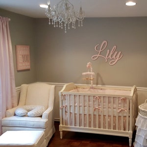 May include: A nursery with a crib, armchair, and ottoman. The crib is white with pink bedding and bows. A mobile hangs above the crib. The name "Lily" is displayed on the wall. A pink curtain and a framed sign that says "Everything about YOU" are also visible.