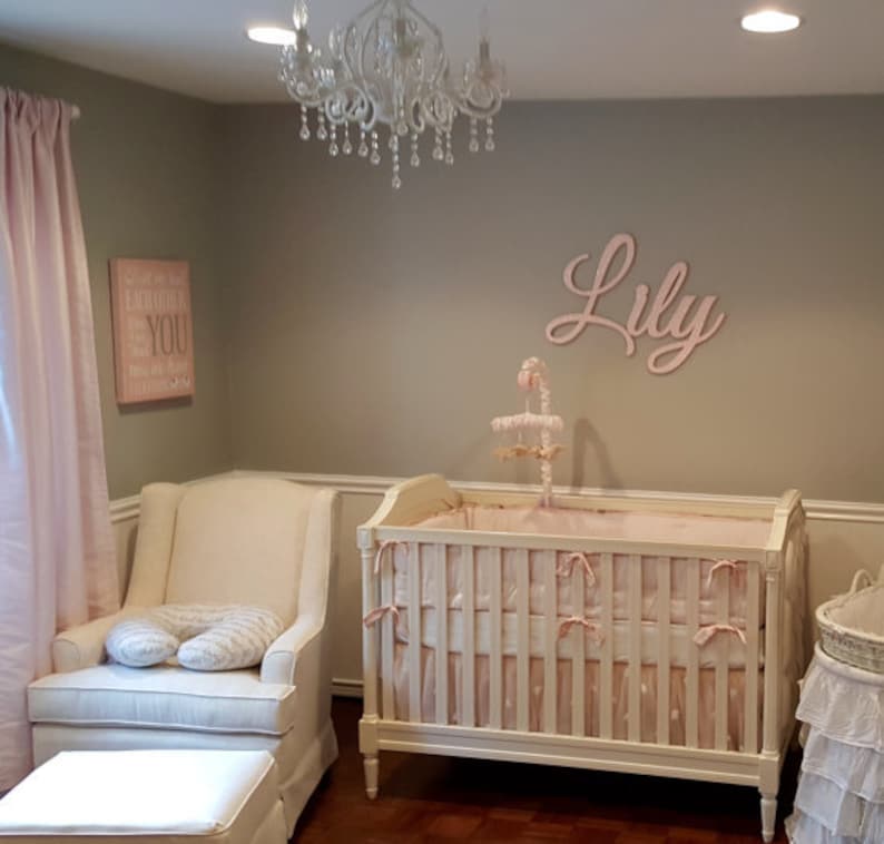 May include: A nursery with a crib, armchair, and ottoman. The crib is white with pink bedding and bows. A mobile hangs above the crib. The name "Lily" is displayed on the wall. A pink curtain and a framed sign that says "Everything about YOU" are also visible.