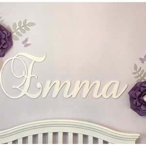 May include: A wall decoration featuring the name "Emma" in white script, surrounded by large paper flowers in shades of purple and white, with gray leaves and butterfly accents. The decor is mounted on a lavender wall.