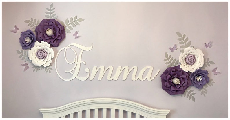 May include: A wall decoration featuring the name "Emma" in white script, surrounded by large paper flowers in shades of purple and white, with gray leaves and butterfly accents. The decor is mounted on a lavender wall.