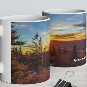 May include: Two white ceramic mugs with a scenic sunset image printed on them. The mug on the left shows a forest scene with trees silhouetted against a colorful sunset sky. The mug on the right shows a mountain range with a sunset sky and the text "Spruce Knob" printed on the bottom right corner.