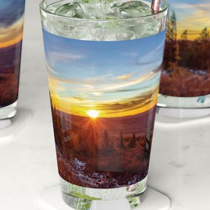 Puede incluir: Tres vasos de vidrio transparentes con imágenes de puestas de sol coloridas impresas en ellos. Cada vaso tiene una pajita y cubitos de hielo. Los vasos están sobre posavasos blancos.