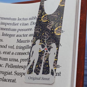 May include: A black metal bookmark with a silhouette of two people standing under a tree. The bookmark has a black and white design with moons, stars, and snakes. The text "Original Smut" is printed on the bookmark.
