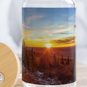 Puede incluir: Una lata de vidrio con una escena de puesta de sol impresa en ella. La lata tiene una tapa de bambú con un agujero para la pajita. La lata está sentada en una mesa de madera.