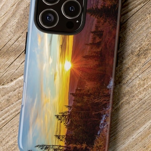 Könnte beinhalten: Eine Handyhülle mit einem Sonnenuntergang über einer Waldszene. Die Hülle hat einen schwarzen Kamerabuckel mit vier Objektiven.