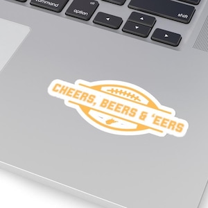 May include: A white sticker with the text "CHEERS, BEERS & 'EERS" in orange and white. The sticker has an oval shape with a football graphic in the center. The sticker is on a laptop.