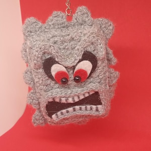 May include: A gray crocheted keychain featuring a Thwomp character from the Super Mario video game series. The Thwomp has a menacing expression with red eyes, a wide open mouth, and a silver keyring.