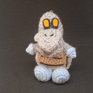 May include: Crocheted Dry Bones character from the Super Mario video game series. The figure is primarily gray with a tan shell and light blue limbs and feet. The eyes are orange.