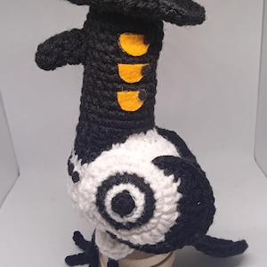 May include: Crocheted bird figure in black, white, and yellow. The bird has a black hat, a long neck with three yellow accents, and a white body with black details. The bird is standing on a small wooden base.