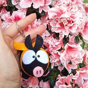 Puede incluir: Un llavero artesanal con un diseño de cerdo de dibujos animados. El cerdo tiene un cuerpo negro, ojos blancos, un hocico rosa y detalles amarillos. El llavero se sostiene frente a un fondo de flores de azalea rosas y blancas.