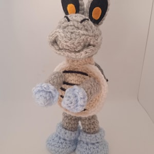 May include: A handmade crochet Koopa Troopa figure. The figure is primarily gray, with a light tan belly and blue accents on the hands and feet. The Koopa Troopa has black eyes and orange inner ears.
