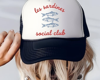 以下が含まれることがあります： 白とネイビーのトラック運転手の帽子。赤い筆記体で「les sardines social club」と書かれ、3匹の青い魚のイラストが描かれています。帽子はメッシュの背面と湾曲したつばが特徴です。