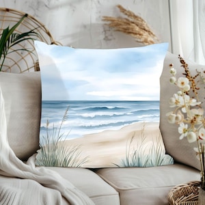 May include: A watercolour pillow featuring a beach scene with blue waves crashing on a sandy shore. The pillow has a light blue background with white clouds and green grass in the foreground.