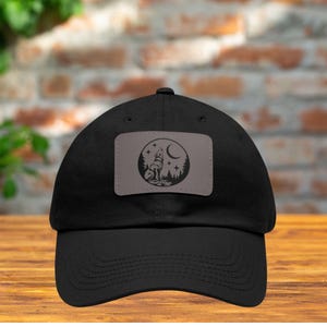 May include: Black baseball cap with a gray patch featuring a howling wolf silhouette under a crescent moon and stars. The patch is surrounded by a forest scene. The cap is made of a durable fabric.
